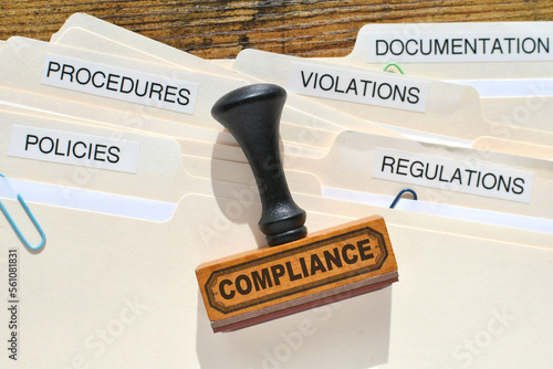 A Compliance rubber stamp on file folders marked Policies Regulations Violations Procedures and Documentation. Compliance in the workplace.