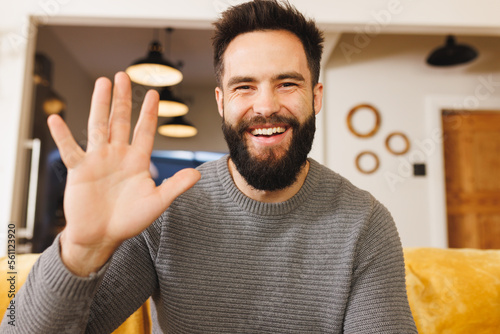 Portrait of happy biracial bearded young man waving hand while sitting on sofa in living room