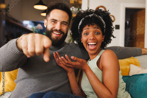 Portrait of cheerful biracial couple laughing and gesturing while sitting on sofa in living room