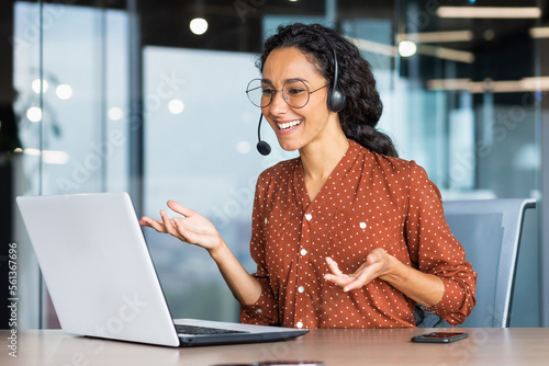 Video call online meeting with colleagues, Hispanic woman working inside modern office, businesswoman smiling and talking remotely using laptop and headset.