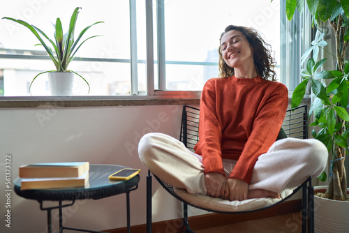 Young relaxed smiling pretty woman relaxing sitting on chair at home. Happy positive beautiful lady feeling joy enjoying wellbeing and lounge chilling near window in modern cozy apartment interior.