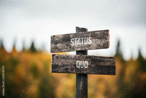 vintage and rustic wooden signpost with the weathered text quote status quo, outdoors in nature. blurred out forest fall colors in the background.