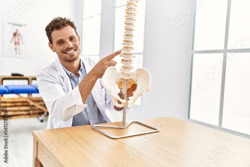 Young hispanic man wearing physiotherapist uniform pointing to anatomical model of vertebral column at clinic