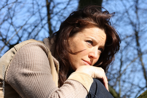 woman leans chin on knees and loking in the sun before background with tree and blue sky