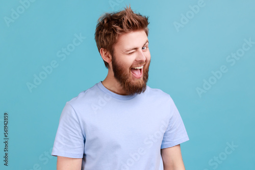 Portrait of bearded man being in good mood, smiling broadly and winking at camera, keeps mouth open, having flirting facial expression. Indoor studio shot isolated on blue background.