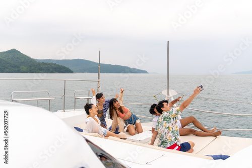 Group of Asian man and woman friends using mobile phone taking selfie together while travel on luxury private catamaran boat yacht sailing in the ocean on summer holiday vacation at tropical island.