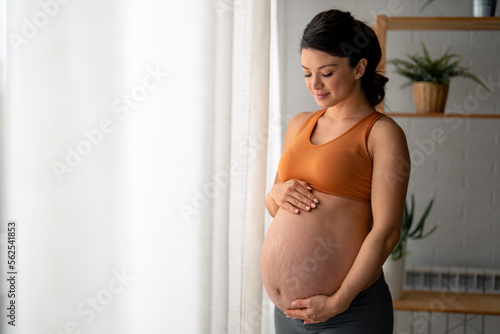 A young expectant mother stroking her baby bump and enjoying her morning, standing near the window at home, wearing comfortable maternity cloths.