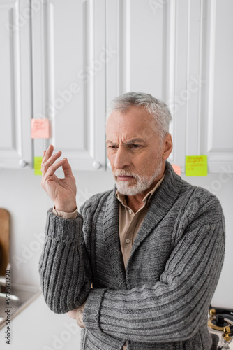 thoughtful man with alzheimer disease standing near blurred sticky notes in kitchen.