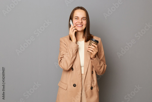 Portrait of cheerful attractive happy woman wearing beige jacket standing isolated over gray background, drinking takeaway coffee and talking on mobile phone, expressing positive emotions.