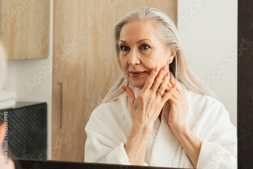 Senior woman with long grey hair touching her cheek with fingers in bathroom