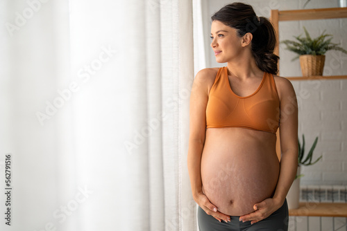 Beautiful pregnant woman touching her belly, standing by the window with the white curtains at home during the day.