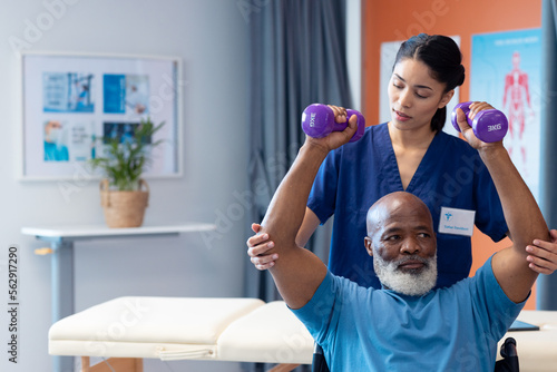 Diverse female physiotherapist helping senior male patient exercise with dumbbells, copy space