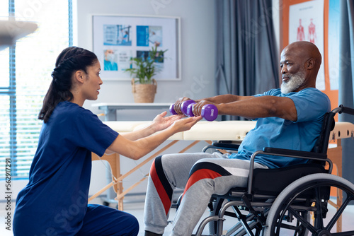Diverse female physiotherapist helping senior male patient in wheelchair exercise with dumbbells