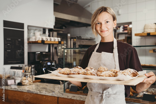 Welcoming female baker holding freshly baked almond croissants in background of bakery shop.