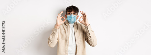 Covid-19, health and real people concept. Excited and impressed guy in sterile face mask showing OK signs in approval, praise cool thing, standing amused on white background