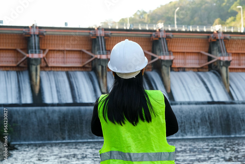 Rear view of female engineer in green vest and helmet standing outside against background of dam with hydroelectric power plant and irrigation.