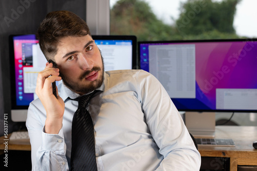 jeune homme d'affaires, ou employé de bureau qui téléphone avec son smartphone, assis devant ses ordinateurs