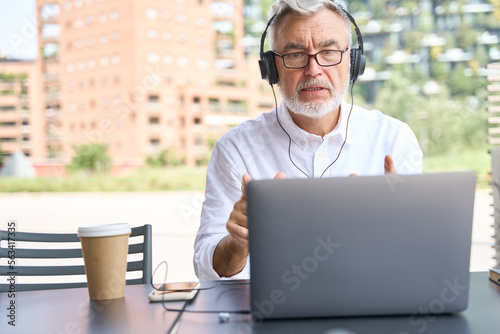 Older senior business man wearing headphones looking at laptop, talking, having hybrid conference online remote video call, virtual distance class or webinar training presentation working outdoors.