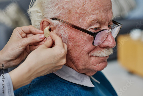 Close-up of senior man in eyeglasses having bad hearing while nurse wearing hearing aid into his ear