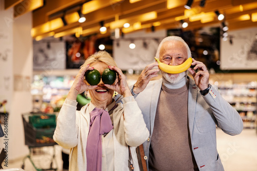 A silly senior couple is pretending fruits are their mouth and eyes while standing at the supermarket.