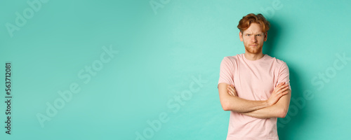 Gloomy redhead guy feeling offended, frowning upser with arms crossed on chest, looking insulted and sulking, standing over mint background