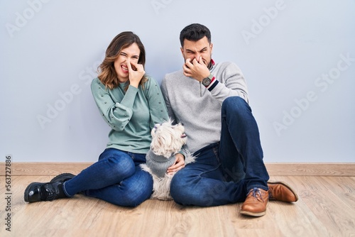 Young hispanic couple sitting on the floor with dog smelling something stinky and disgusting, intolerable smell, holding breath with fingers on nose. bad smell