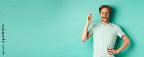 Amused young man with red hair, wearing glasses and t-shirt, showing okay sign and smiling excited, checking out something and approving it, standing over turquoise background