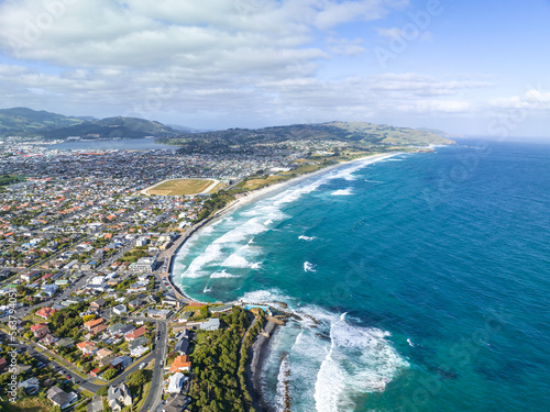 High angle aerial drone view of St Clair, a beachside suburb of Dunedin, second-largest city in the South Island of New Zealand. Dunedin city in the background. Hot water salt rock pool in foreground.