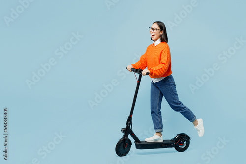 Full body side view cool happy young woman of Asian ethnicity wear orange sweater glasses riding electric scooter isolated on plain pastel light blue cyan background studio People lifestyle concept.