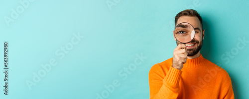 Funny man looking through magnifying glass, searching or investigating something, standing in orange sweater against turquoise background