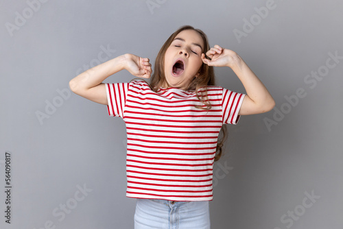 Portrait of sleepy little girl wearing striped T-shirt yawning and raising hands up, feeling fatigued, standing with close eyes. Indoor studio shot isolated on gray background.
