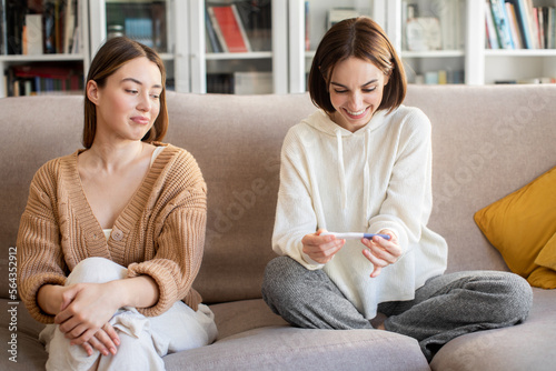 Smiling displeased european millennial lady looking at happy surprised woman with pregnancy test