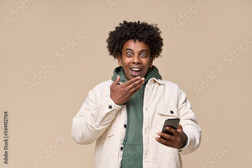 Excited surprised African teen guy holding cell phone receiving message with wow promotion. Amazed ethnic teenager student boy winner using mobile feeling shocked isolated on beige background.