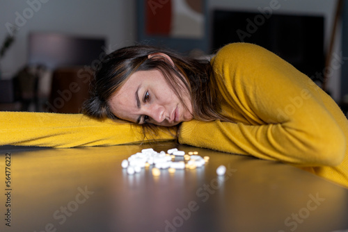 Adult melancholic female with long dark hair in yellow sweater leaning head on hand at table with pile of antidepressant pills at home