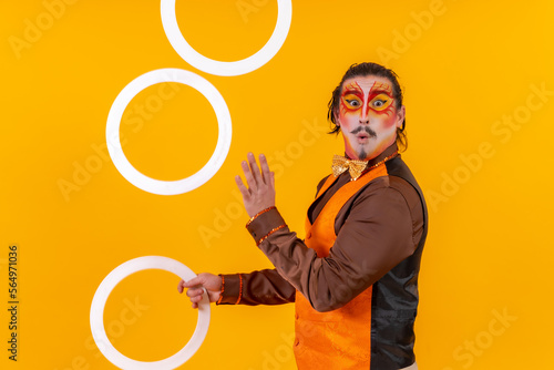Portrait of a juggler in a vest with a painted face juggling hoops on a yellow background