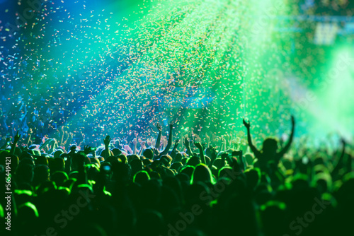 A crowded concert hall with scene stage green lights, rock show performance, with people silhouette, colourful confetti explosion fired on dance floor air during a concert festival