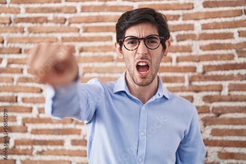 Young hispanic man standing over brick wall background pointing displeased and frustrated to the camera, angry and furious with you
