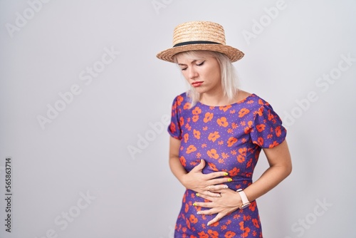 Young caucasian woman wearing flowers dress and summer hat with hand on stomach because indigestion, painful illness feeling unwell. ache concept.