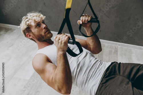 Sideways young concentrated strong sporty athletic sportsman man wear white tank shirt black shorts doing exercises with trx loops looking camera warm up training indoor at gym. Workout sport concept.