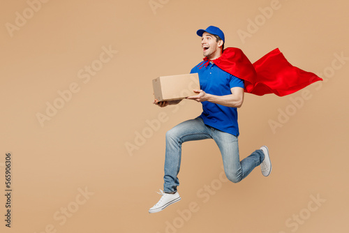 Full body delivery guy employee man wear blue cap t-shirt uniform workwear super hero red coat work as dealer courier hold cardboard box jump high isolated on plain beige background. Service concept.