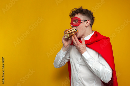 young guy in a superhero costume eats a delicious burger on a yellow background, super man eats fast food