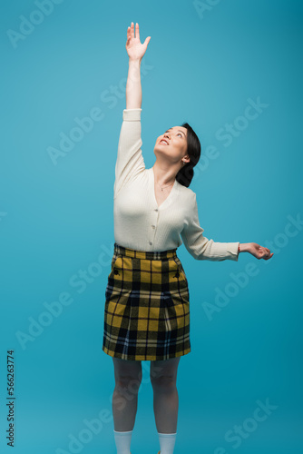 young asian woman in plaid skirt standing with outstretched hand and looking up isolated on blue