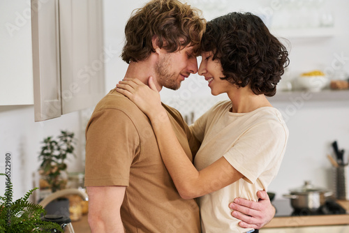 Side view of young amorous husband and wife touching by their foreheads and noses while woman keeping hands on shoulders of her husband