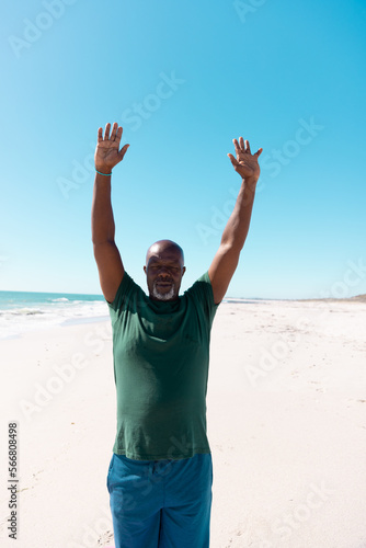 African american bald senior man with arms raised meditating on sandy beach under clear sky