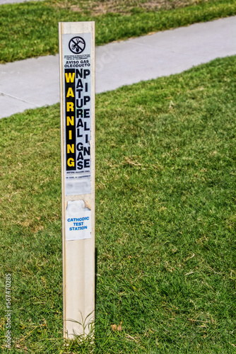 Marker for natural gas pipeline - cathodic test station stuck into green grass by sidewalk - Warning in English and Spanish