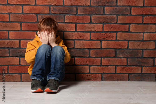 Upset boy sitting on floor near brick wall, space for text. Children's bullying