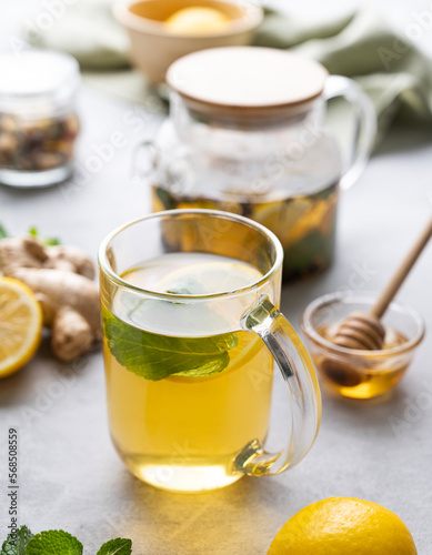 A glass of tea with fresh lemon, honey, mint and ginger on a light background with teapot and morning light close up.