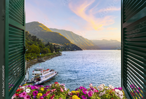 View from a balcony terrace with flowers and shutters of the mountains and lakefront promenade on Lake Como at the town of Bellagio, Italy, under a colorful sunset.