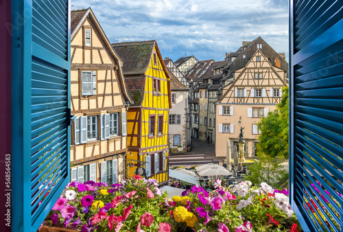 View from an open window overlooking a small square with cafes and half timber frame buildings in the medieval historic center of Colmar, France.