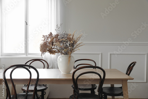Elegant dining room, scandinavian interior. Vase with dry flowers on wooden table. Vintage chairs. White wall moulding background, stucco decor and window. Empty copy space. Elegant flat, home.